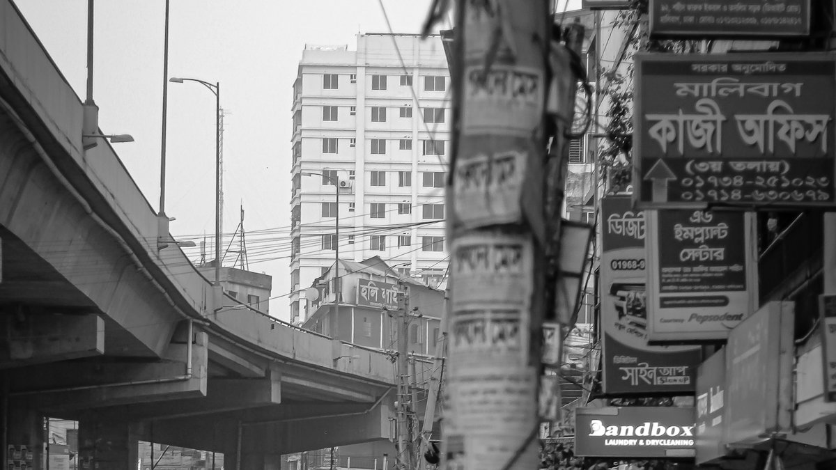 A Dhaka street in black and white, shop signs in Bangla stacked above a flyover
