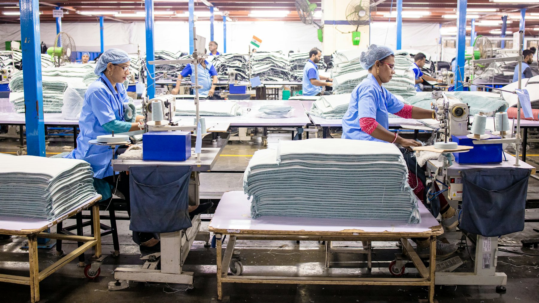 Machinists at rows of sewing tables on a garment factory floor, stacks of cut cloth beside each station