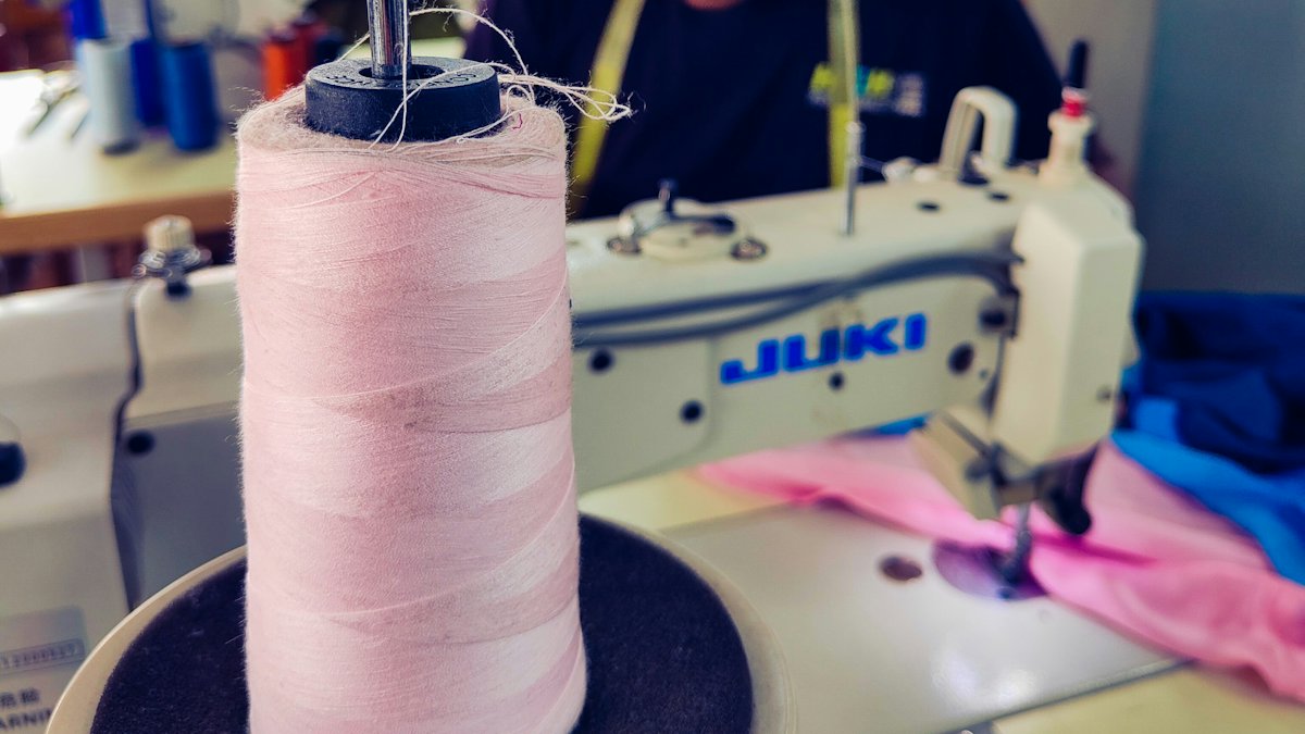 A cone of pink thread mounted on an industrial sewing machine, fabric under the needle