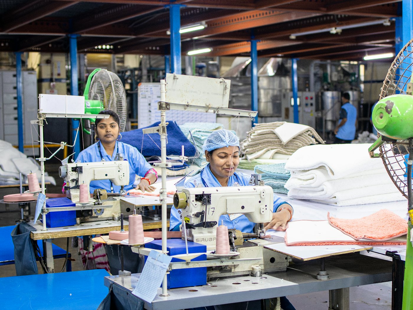 Two machinists at a sewing line, folded towels stacked on the table beside them